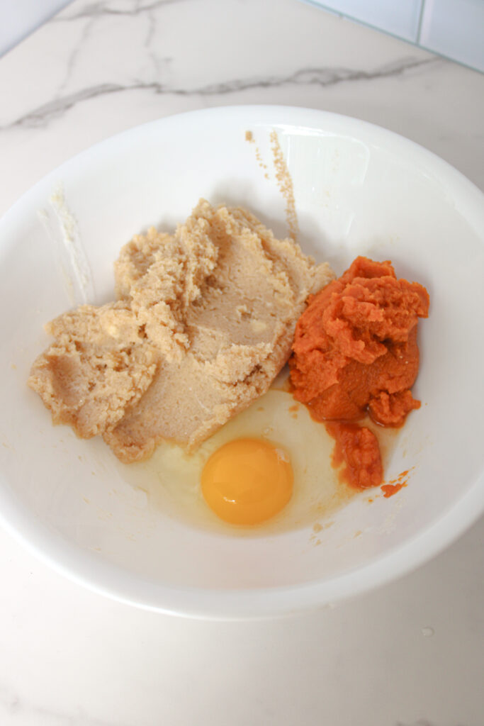 Pumpkin puree and an egg added to creamed butter and sugar in a white bowl.