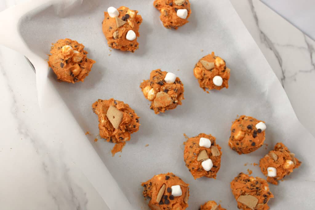 Scoops of pumpkin s’mores cookie dough with marshmallows and graham crackers on a baking sheet.