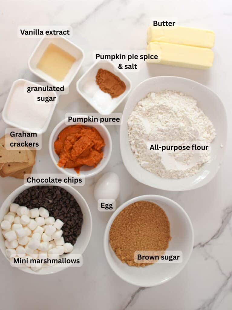 Ingredients for pumpkin s’mores cookies arranged in bowls on a marble surface.