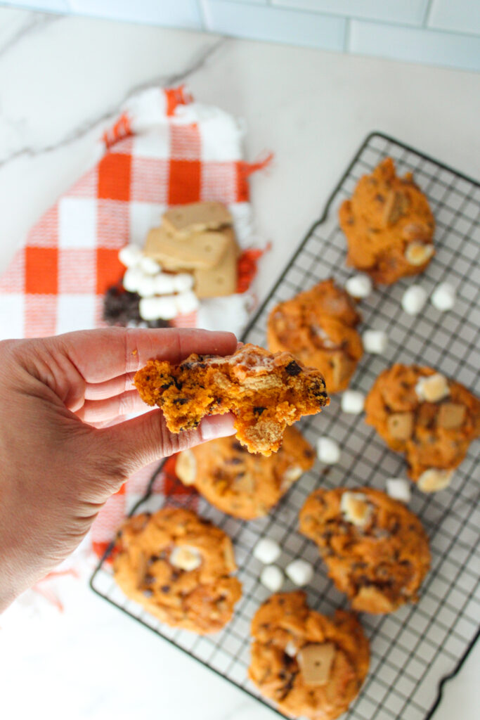 Hand holding a pumpkin s’mores cookie showing its soft, gooey center above a cooling rack.