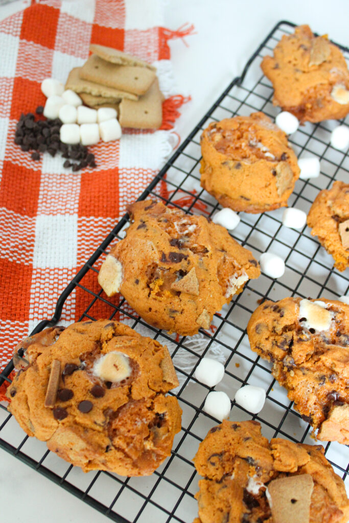 Freshly baked pumpkin s’mores cookies cooling on a wire rack with marshmallows and chocolate chips.