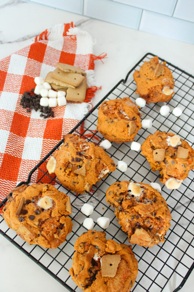 Pumpkin s’mores cookies cooling on a wire rack with marshmallows and graham crackers nearby.